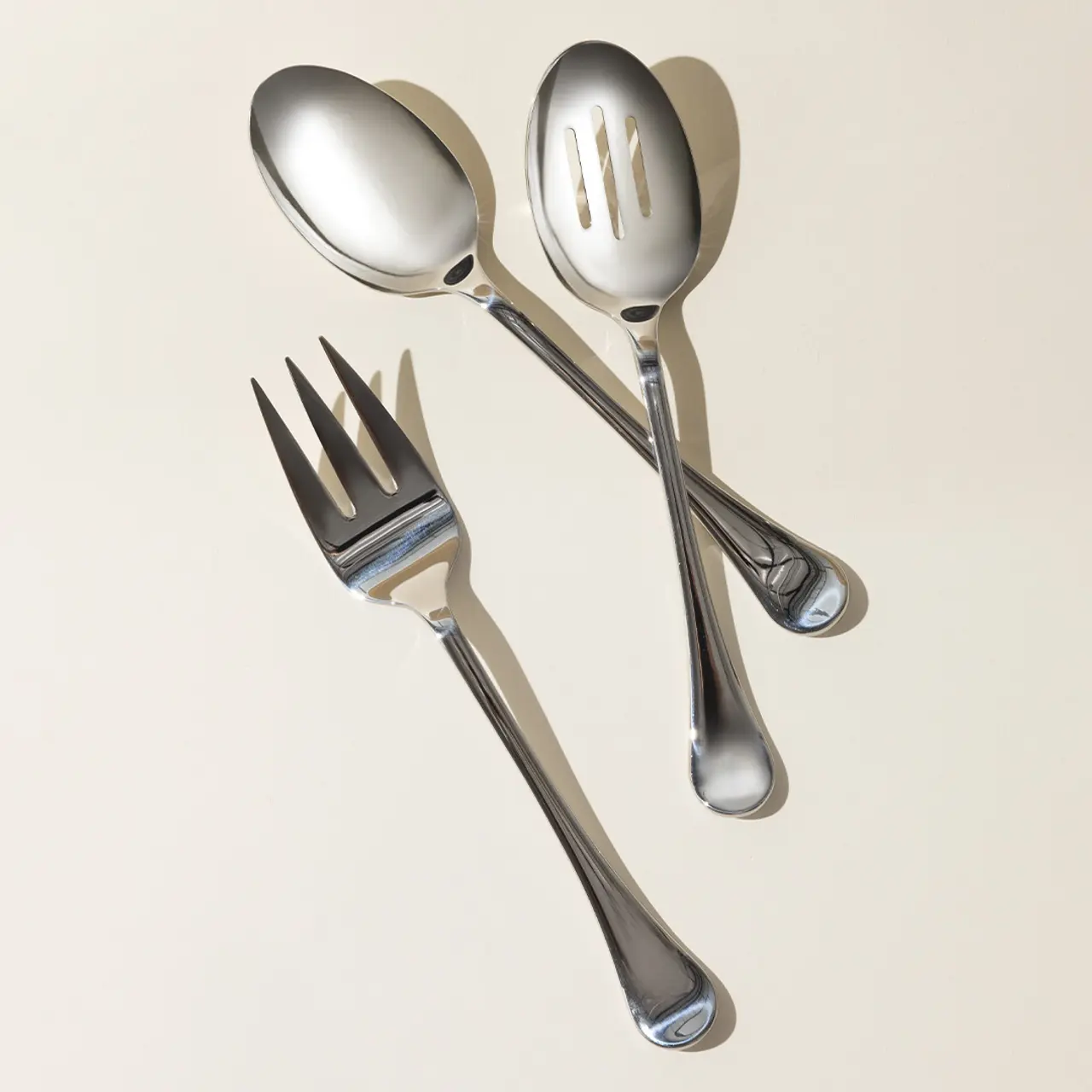 A set of stainless steel cutlery consisting of a fork, a spoon, and a slotted spoon arranged on a neutral background.