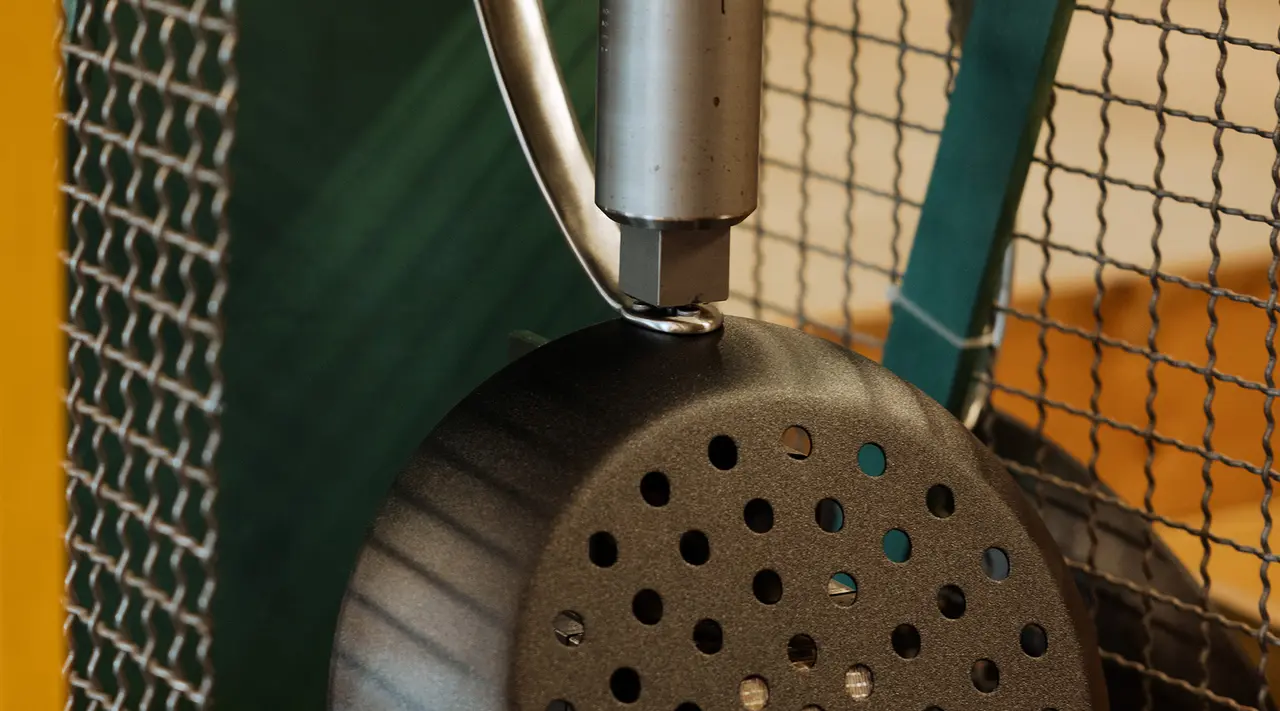 A metal spaghetti strainer with a black handle hangs against a blurred background of a yellow wall and wire mesh.