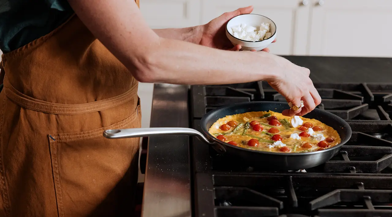 A person in an apron is adding ingredients to a colorful omelet on a stove.