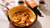 A delicious Dutch baby pancake topped with caramelized apples is served in a skillet, accompanied by two cups of coffee and a bag of coffee beans.