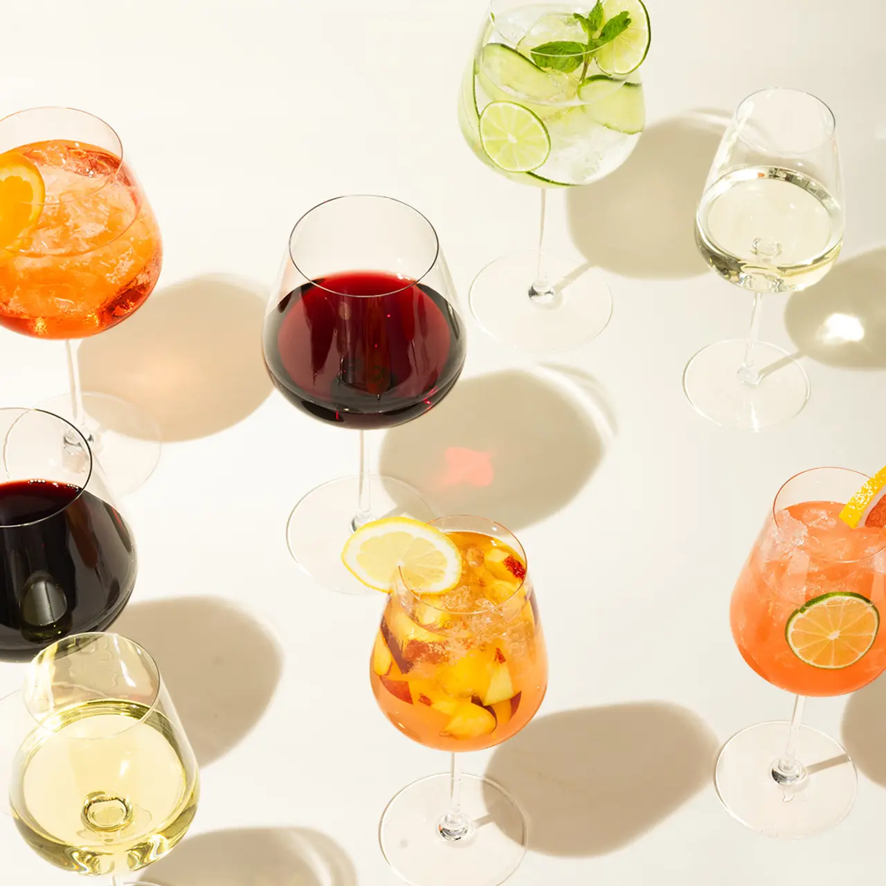 Various glasses of colorful beverages, including wine and cocktails, are arranged neatly, casting shadows on a bright surface.