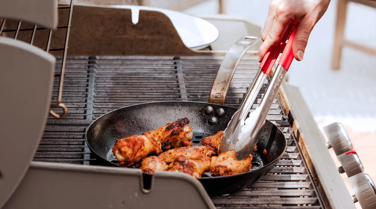 A person is grilling chicken in a skillet on an outdoor grill using tongs.