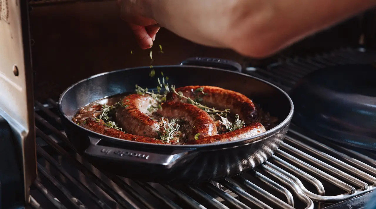 A person seasons sausages sizzling in a pan on a stove, sprinkling herbs for added flavor.