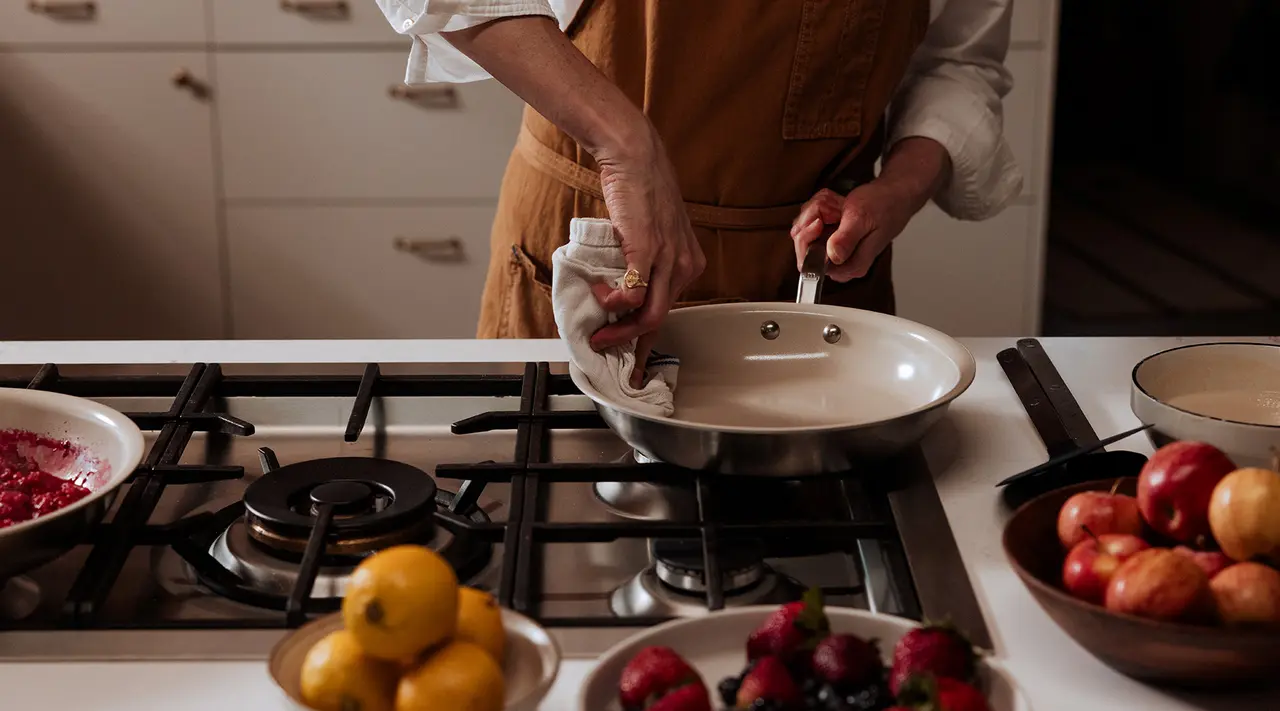 A person in an apron is holding a pan over the stove in a kitchen with fresh fruits and vegetables on the counter.