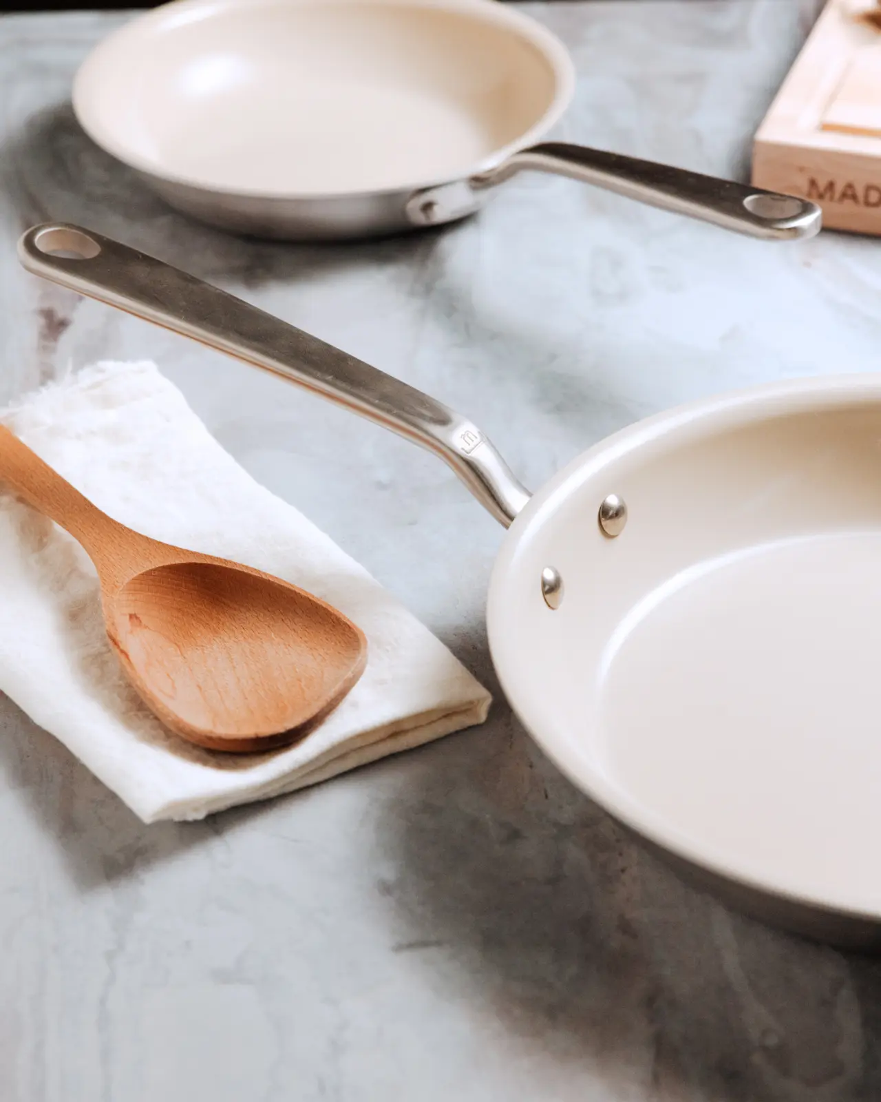 Two empty frying pans and a wooden spoon rest on a marbled countertop, suggesting preparation for cooking.