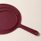 A stack of maroon non-stick frying pans with a handle lies against a neutral background.