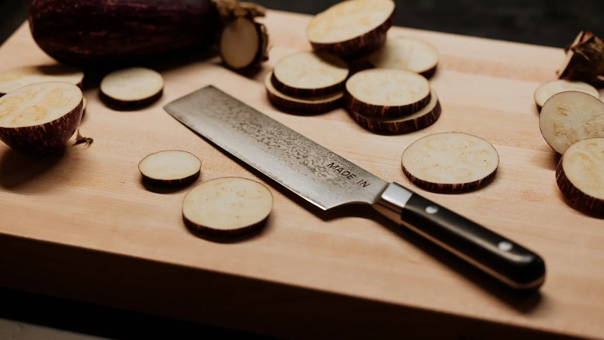 Sliced eggplant pieces are scattered on a wooden cutting board next to a large kitchen knife.