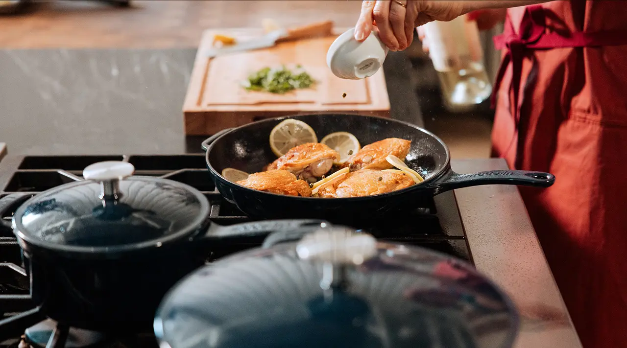 A person in an apron sprinkles seasoning onto chicken fillets cooking in a skillet on the stove, with chopped herbs in the background.