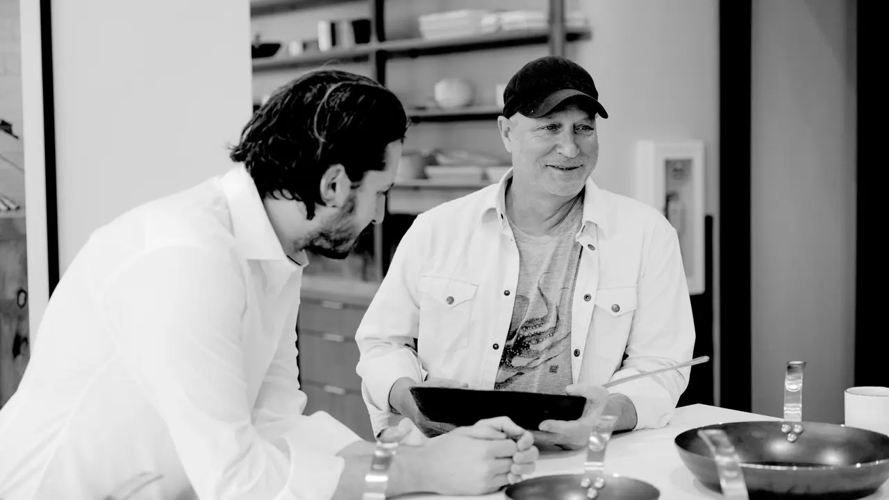 Two men, one holding a tablet, stand in a kitchen with bowls and utensils on the counter, engaged in a pleasant conversation.