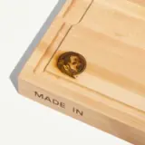 A wooden cutting board features a carved side with a portrait and the text "MADE IN."