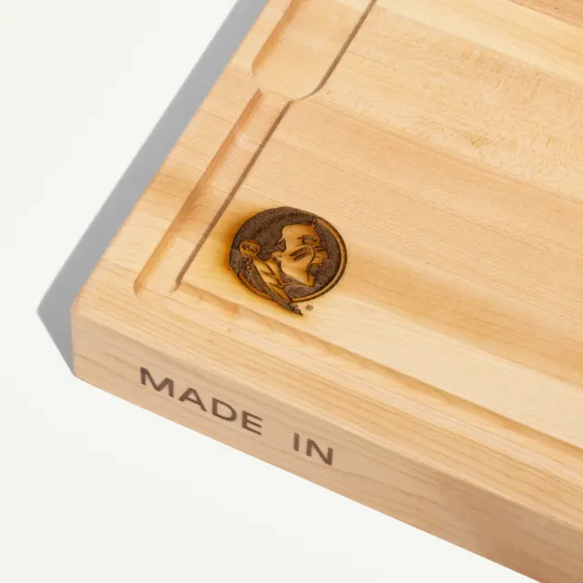 A wooden cutting board features a carved side with a portrait and the text "MADE IN."