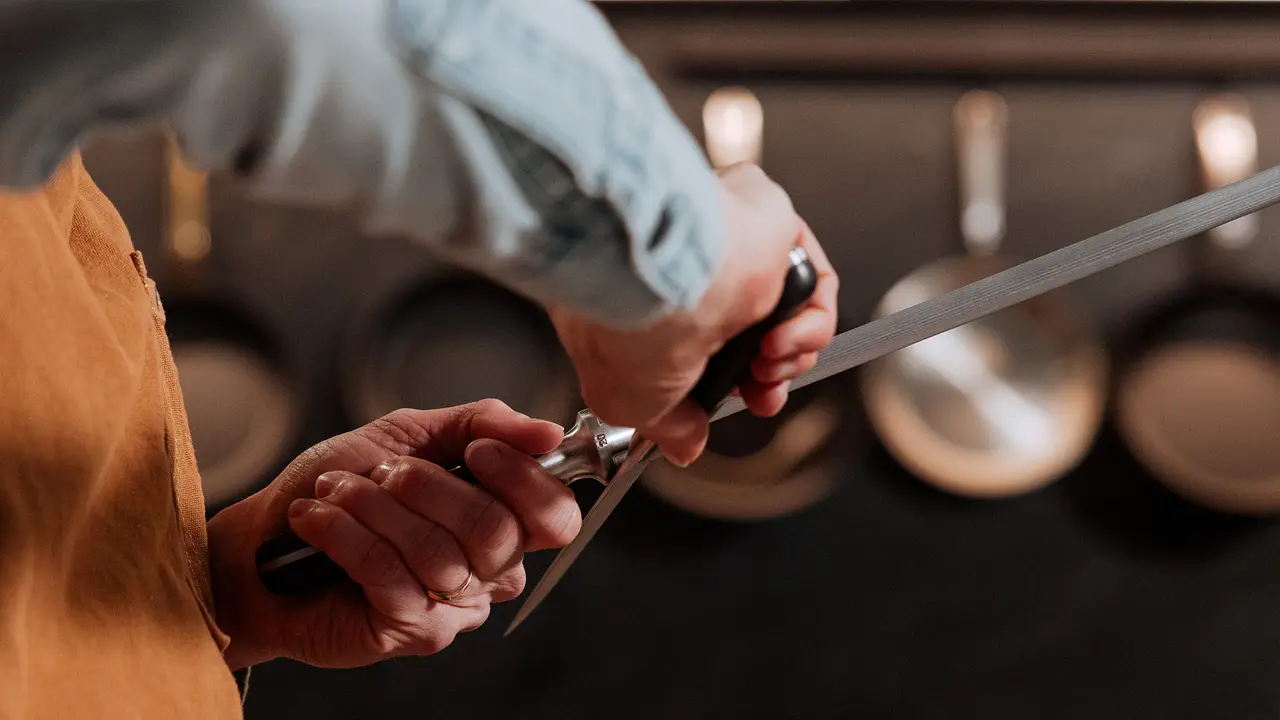 A person sharpens a knife with a honing steel in a kitchen setting.