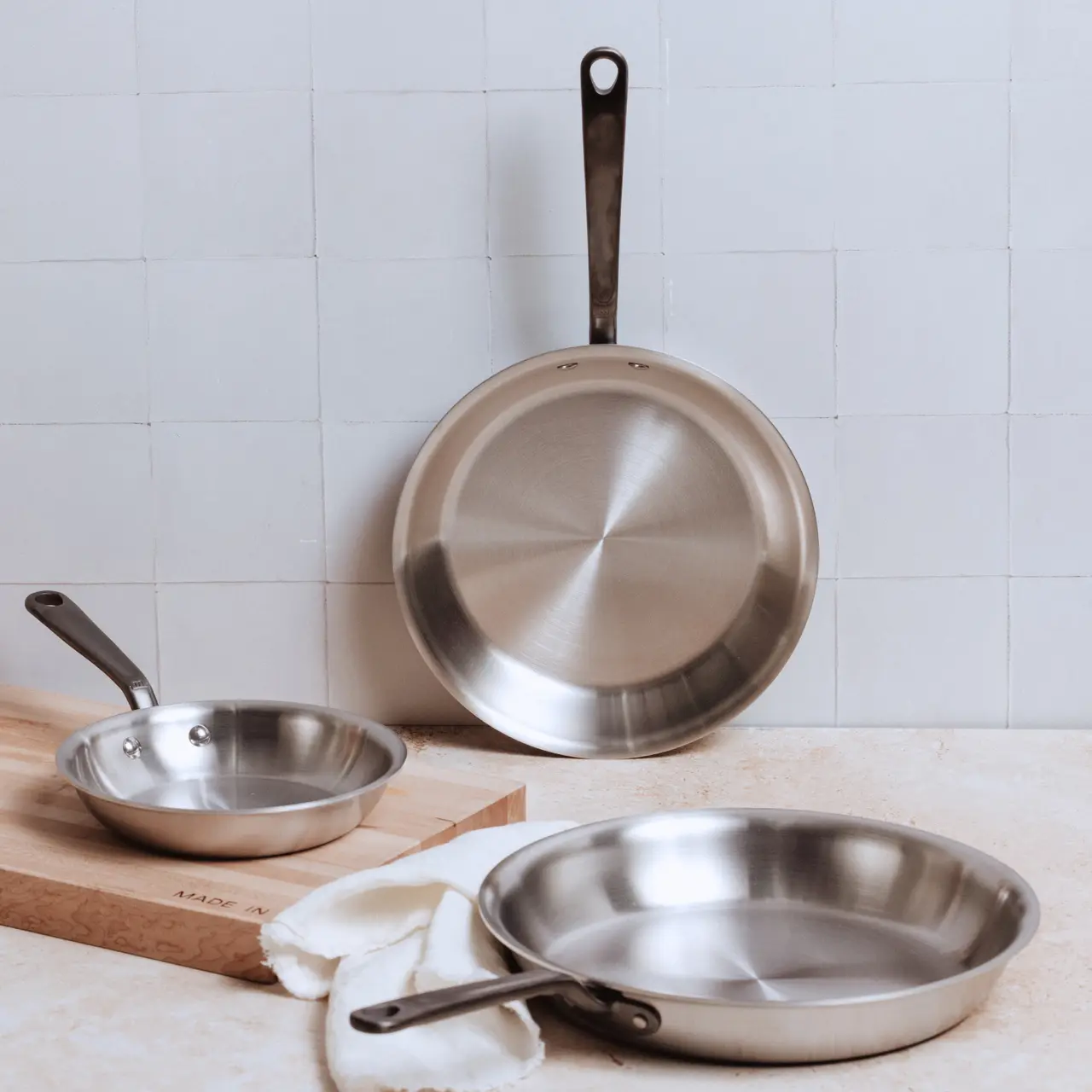 Three stainless steel frying pans of varying sizes are arranged on a kitchen counter with one hanging on the wall.