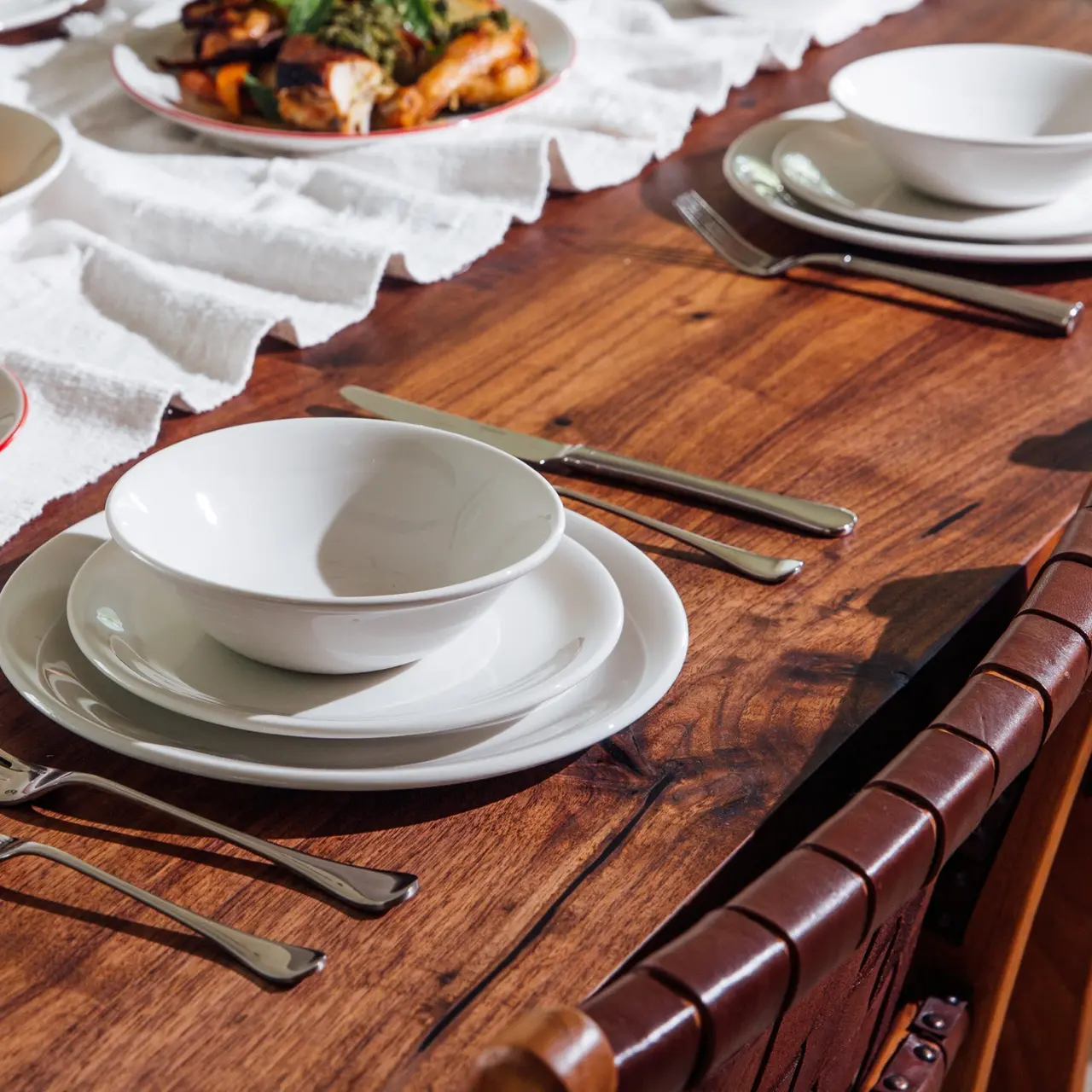 A wooden dining table set with white plates, bowls, and silverware, with a plate of food visible in the background.