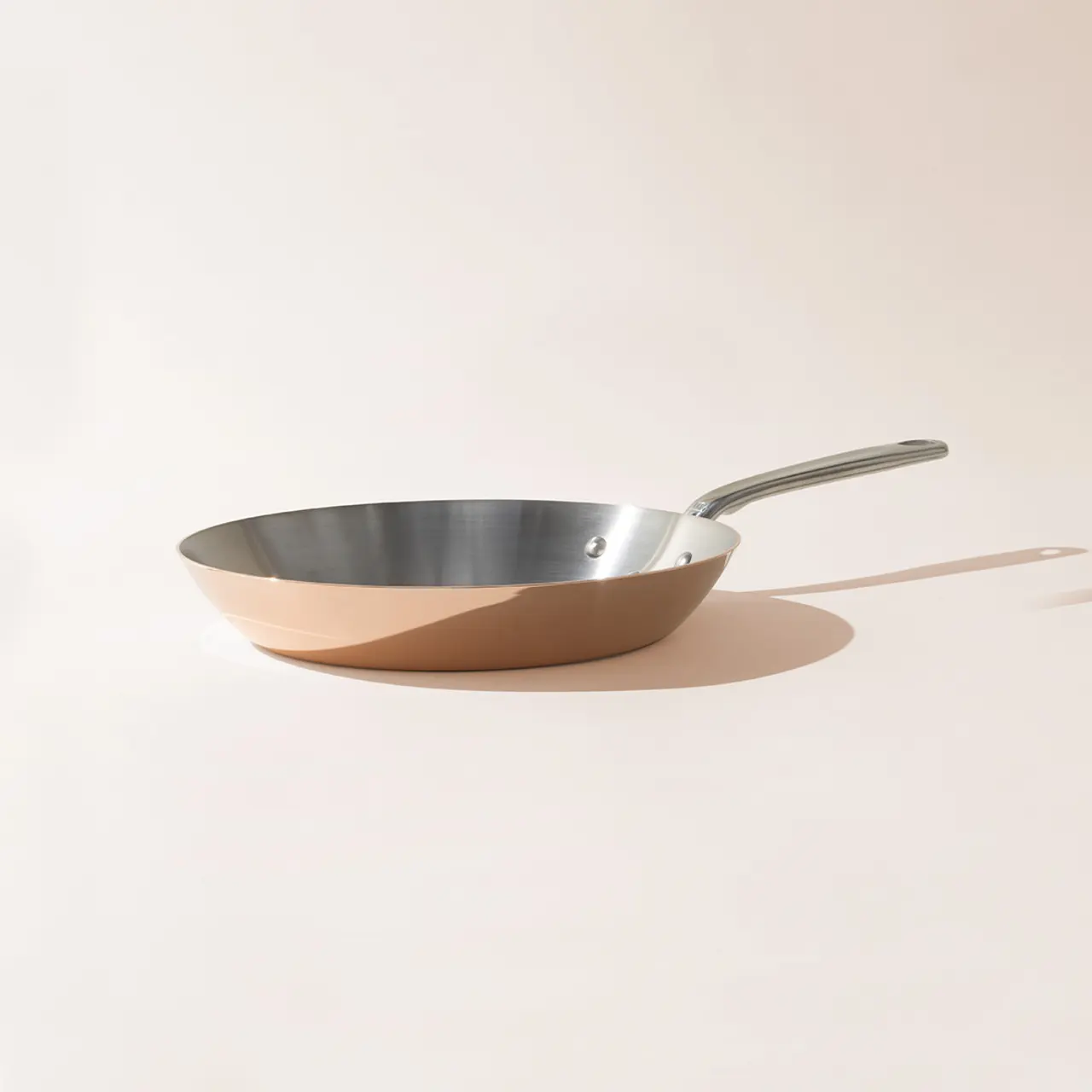 A stainless steel frying pan with a long handle is positioned against a light background, casting a sharp shadow.