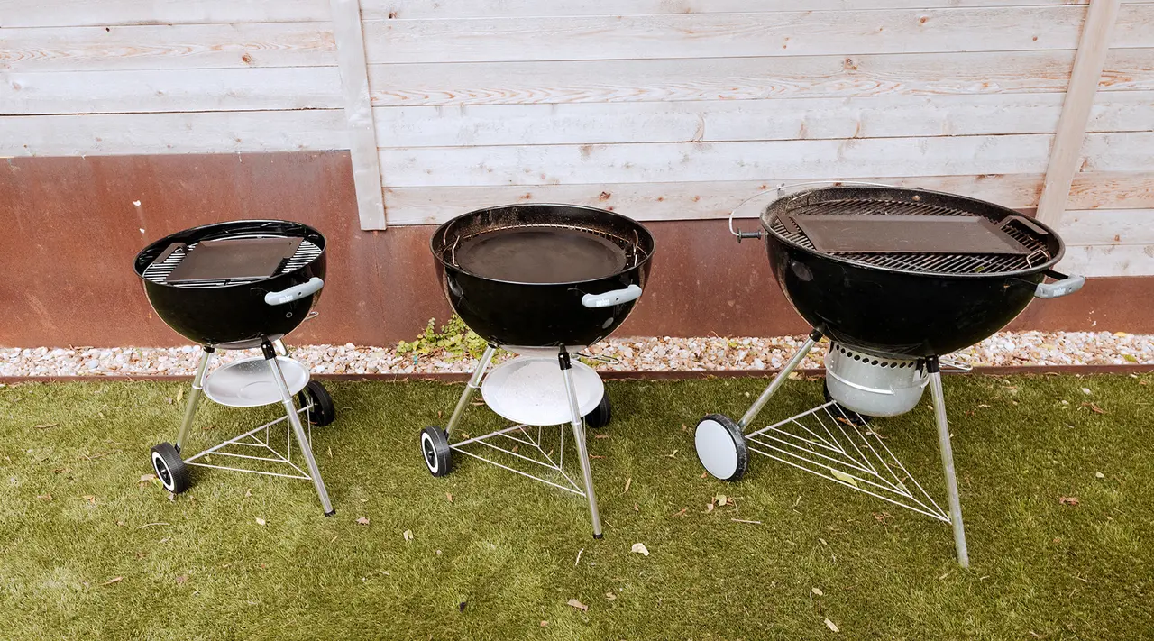 Three black charcoal grills of varying sizes are arranged in a row on a grassy surface against a wooden fence backdrop.