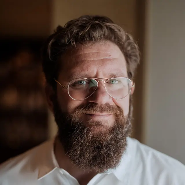 A portrait of a smiling man with a full beard and glasses, wearing a white shirt against a blurred background.