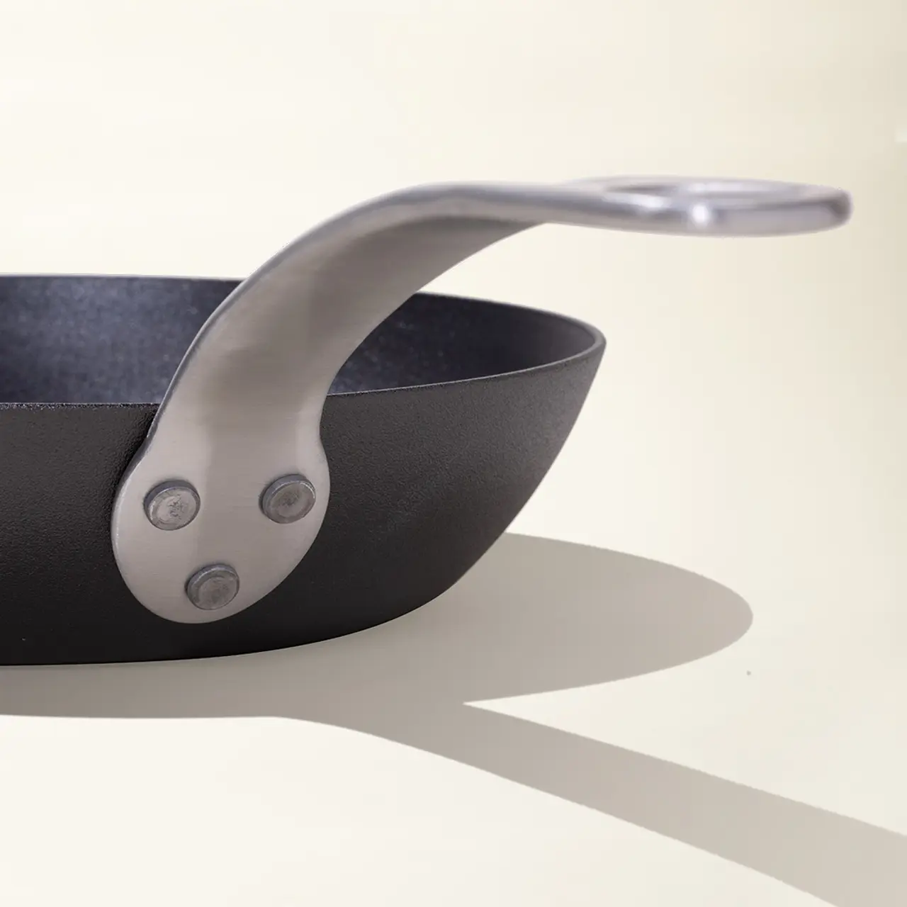A close-up of a pan's handle, showing a sleek metal design with rivet details and a soft shadow cast on a light background.