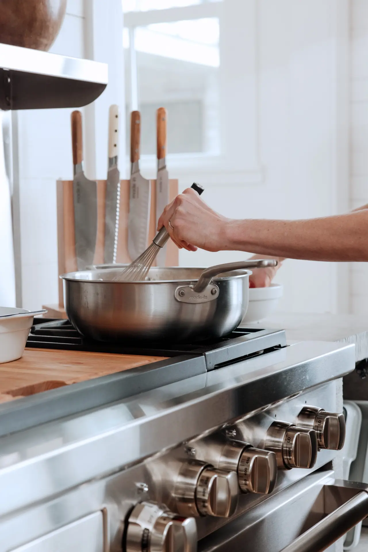 A hand holds a whisk, stirring a pot on a modern stovetop, with knives displayed in the background.