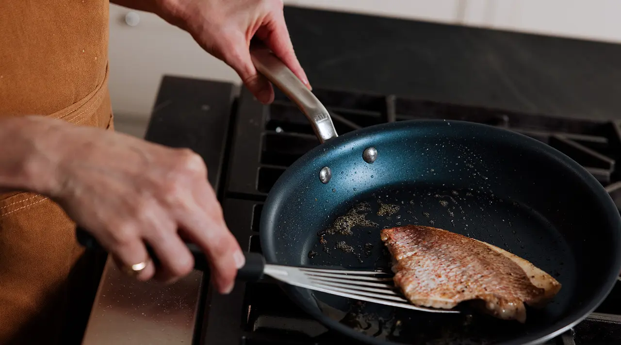 A person is cooking a piece of meat on a skillet over a gas stove.