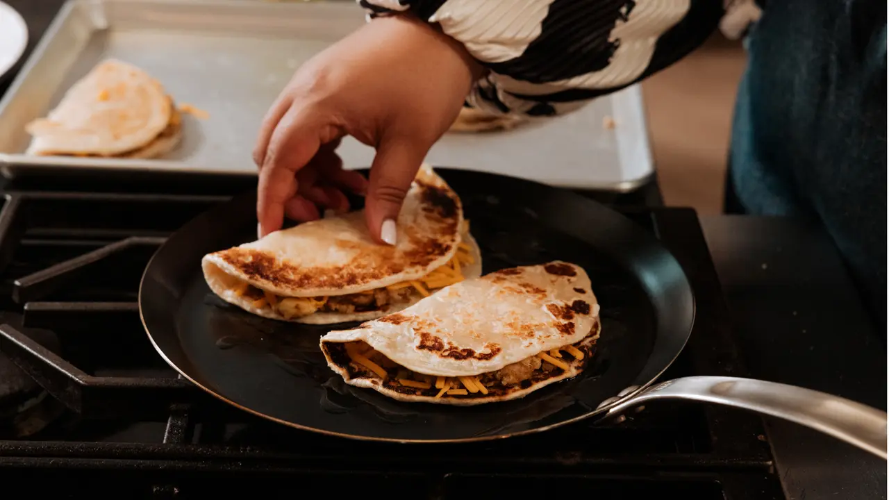 A person is flipping quesadillas on a black pan over a stovetop, showing a cooking process in action.