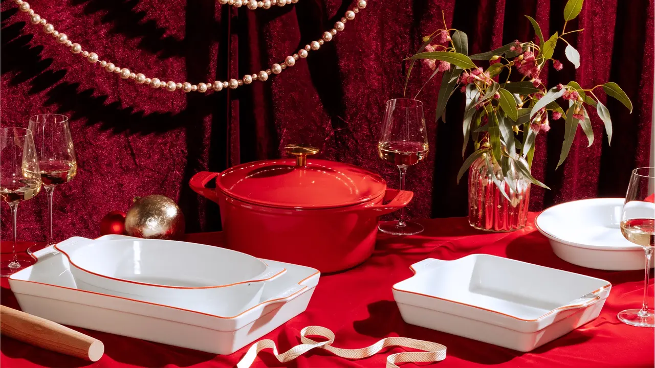 Festive table setting featuring elegant cookware and serveware with a red and white theme, accompanied by wine glasses and holiday decorations on a red backdrop.