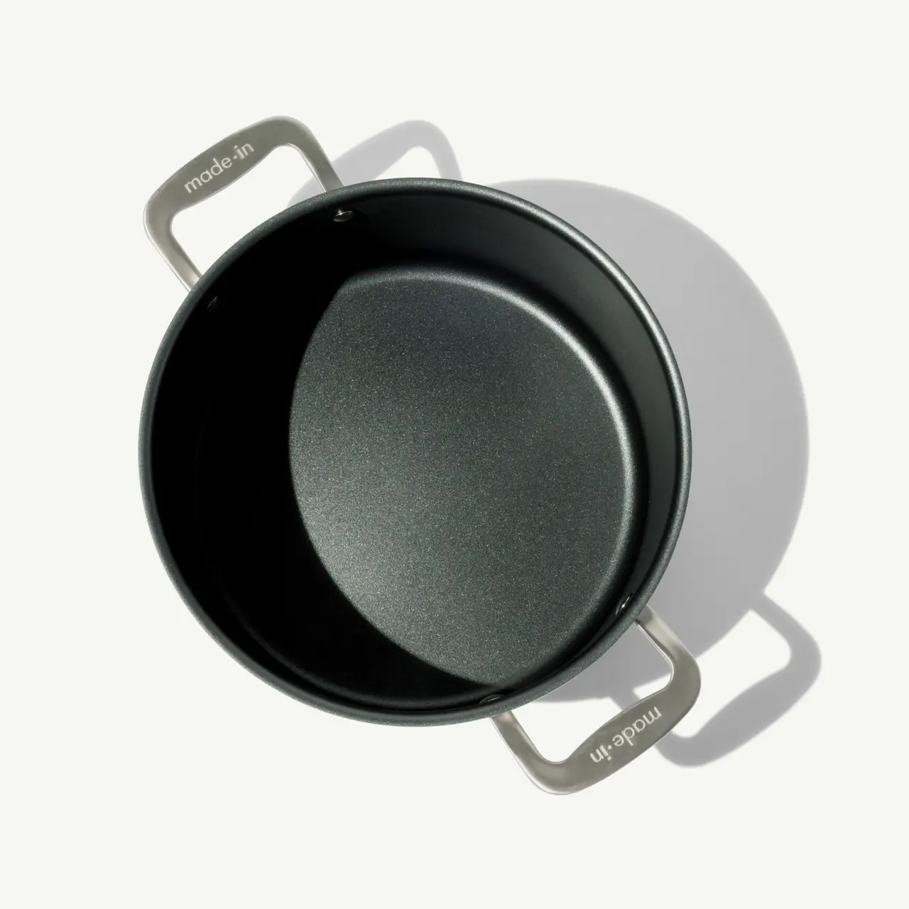 A black non-stick frying pan with two silver handles on a light background casting a distinct shadow.