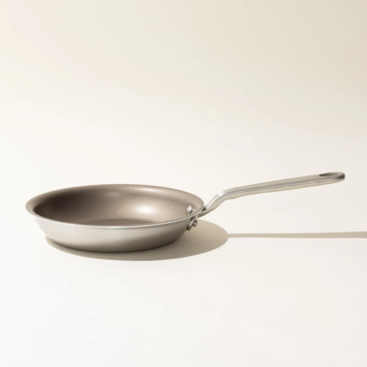 A stainless steel frying pan with a long handle is positioned to the left on a light background with a noticeable shadow to its right.