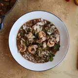 A white bowl filled with rice, shrimp, chicken, and greens in a savory broth is centered on a textured surface.