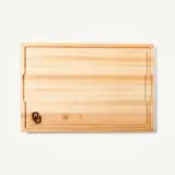 A wooden cutting board features a smooth surface and a logo in the bottom corner.