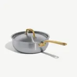 A silver saucepan with a gold handle and lid is positioned on a light background.