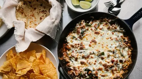 A skillet of cheesy baked dish with green peppers is served alongside tortilla chips, fresh lime wedges, and warm tortillas.