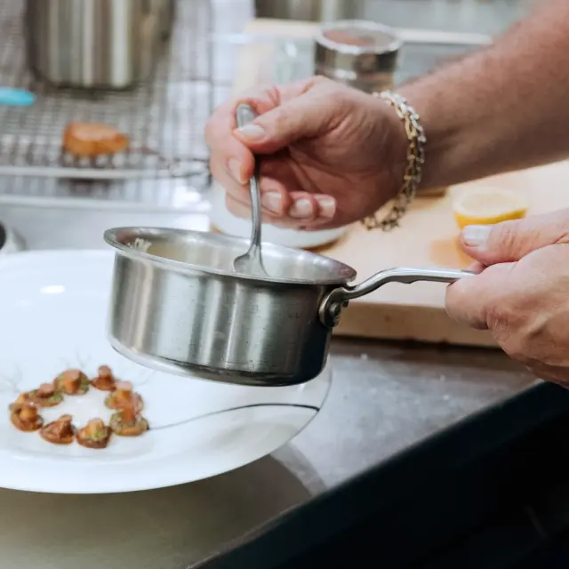 A person with a bracelet is stirring sauce in a small saucepan while holding a spoon, with a garnished plate of food nearby.