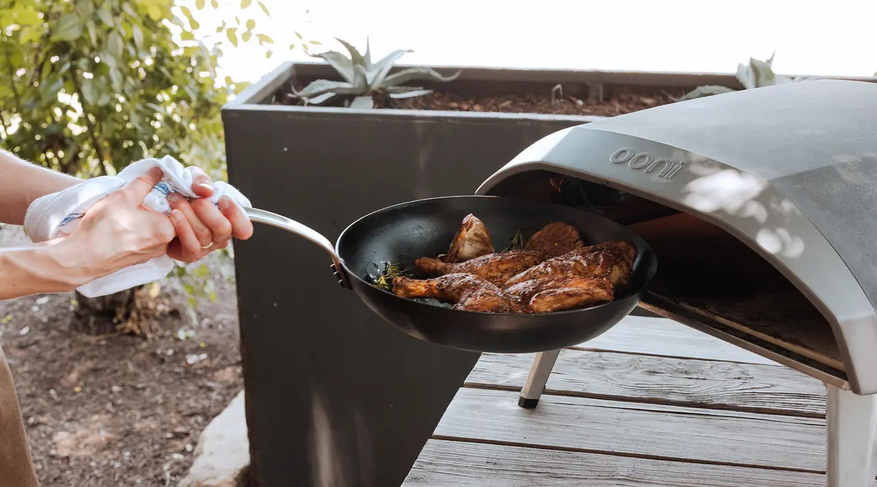 A person is pulling out a pan of grilled chicken from an open outdoor grill oven.