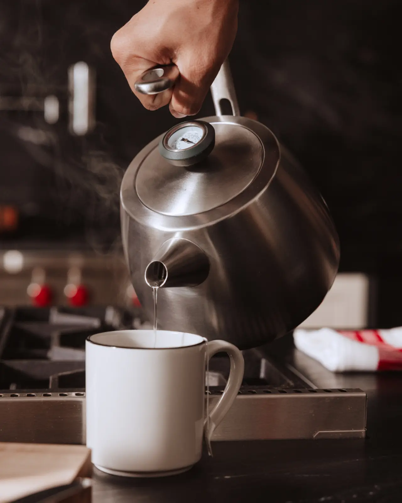 A hand pours steaming water from a stainless steel kettle into a white mug on a kitchen countertop.