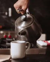 A hand pours steaming water from a stainless steel kettle into a white mug on a kitchen countertop.