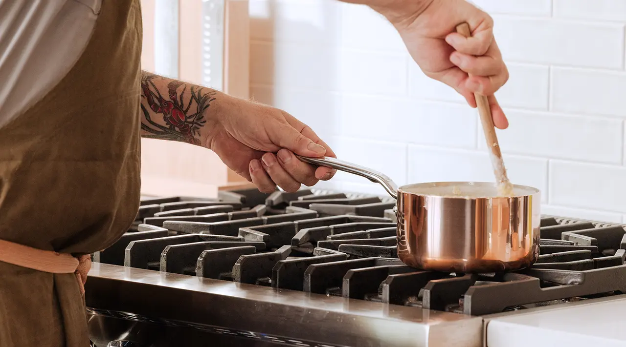 A person with a tattoo on their arm is stirring something in a copper pot on a gas stove.