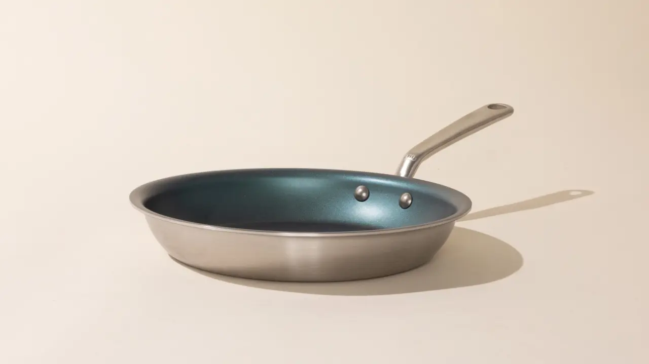 A stainless steel frying pan with a non-stick coating and three rivets attaching the handle, casting a slight shadow on a plain surface.