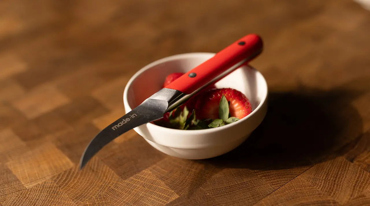 A red paring knife rests on a white bowl containing sliced strawberries on a wooden table.