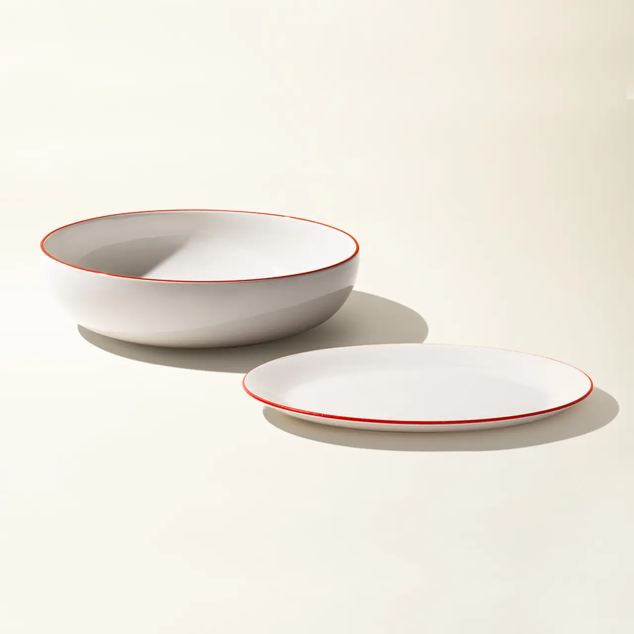 A white bowl and plate with a red rim are neatly placed against a cream background.