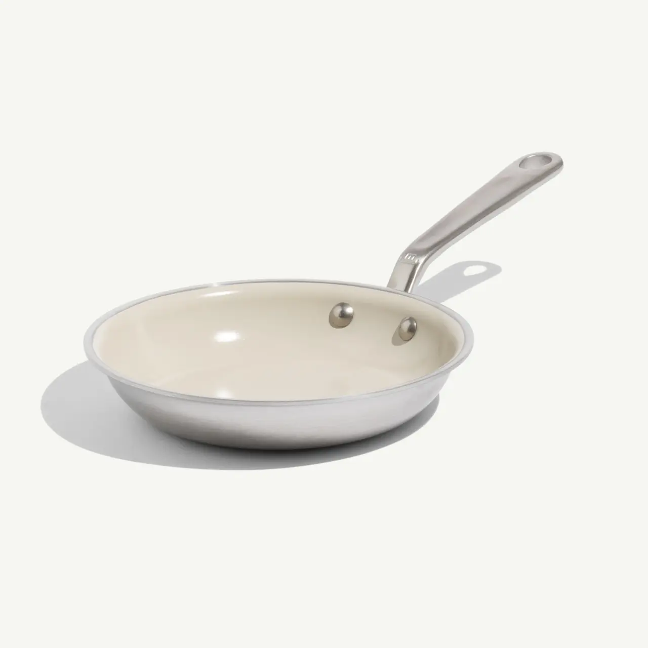 A stainless steel frying pan with a beige interior and a long handle is placed on a light background.