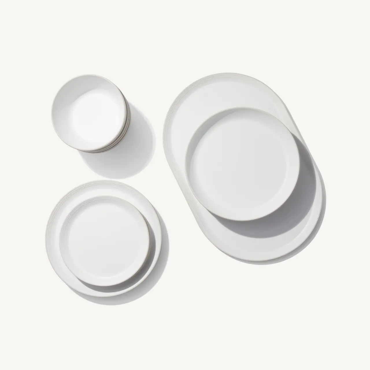 White ceramic dishware, consisting of stacked round plates and an oval platter, is neatly arranged on a light surface casting soft shadows.
