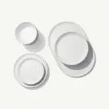 White ceramic dishware, consisting of stacked round plates and an oval platter, is neatly arranged on a light surface casting soft shadows.