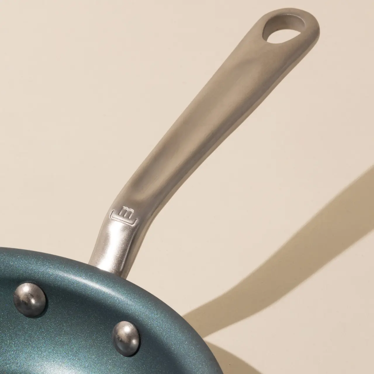A close-up of a teal frying pan with a silver handle against a light background with a shadow stripe across it.