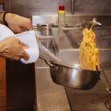 A hand holds a pan over a stovetop as noodles and sauce flambé dramatically rise from it.