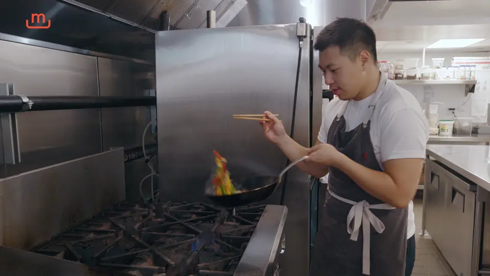 A chef is focused on stir-frying food in a wok over an open flame in a commercial kitchen.