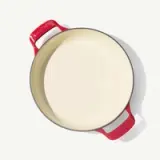 A round, red-handled cast iron pot with a smooth, light-colored interior sits on a neutral background.