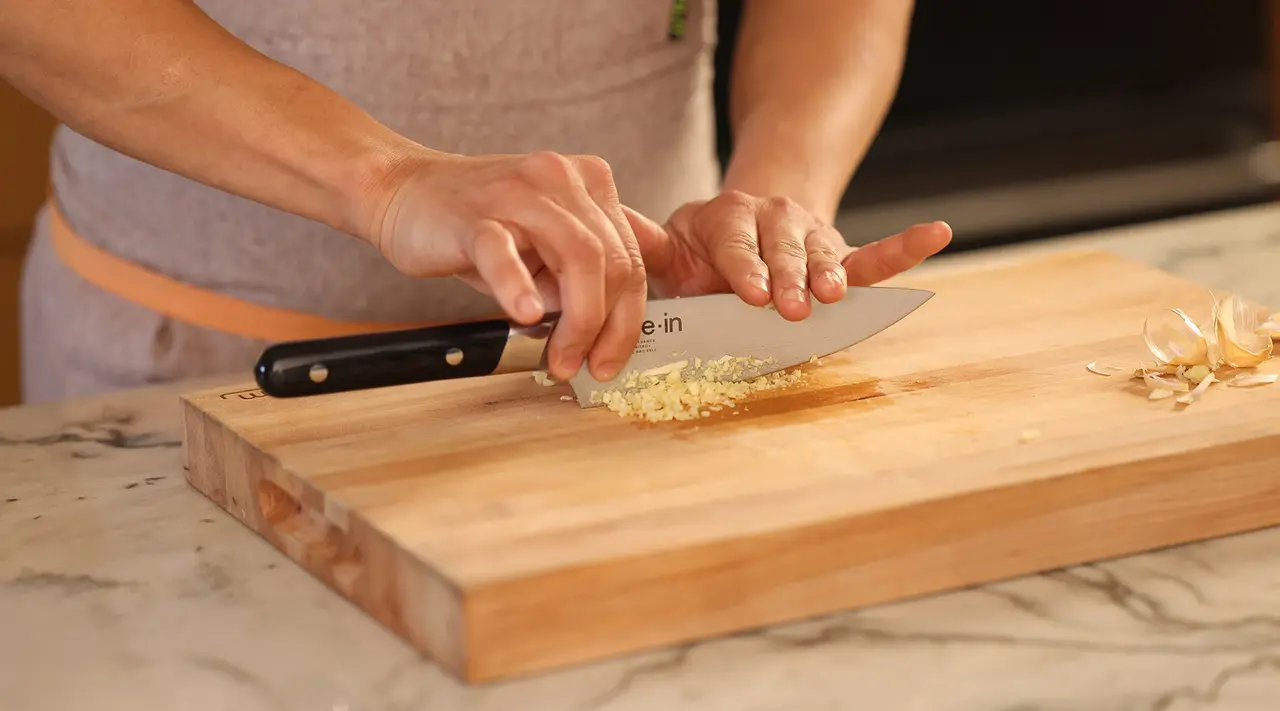 A person is mincing garlic on a wooden cutting board using a chef's knife.