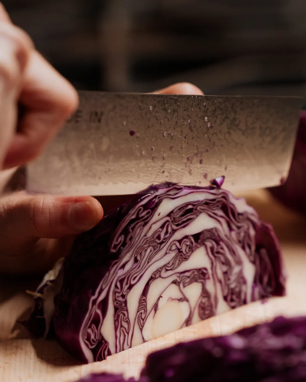 A person's hands are slicing a red cabbage with a knife on a wooden cutting board.