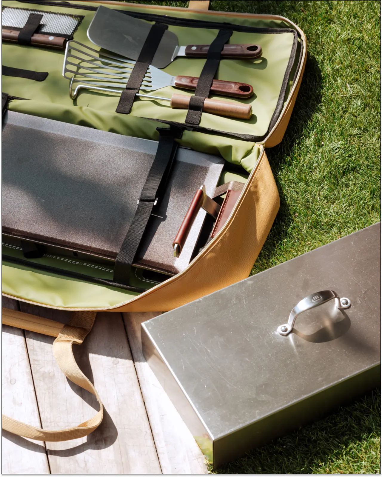 A barbecue tool set is displayed open on the grass, showcasing various grilling utensils next to a closed metal case.